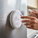 Hands polishing a lightly scratched stainless steel kitchen appliance with a microfiber pad and polishing compound bottle.