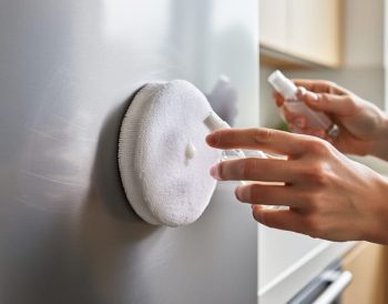 Hands polishing a lightly scratched stainless steel kitchen appliance with a microfiber pad and polishing compound bottle.