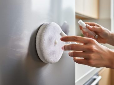 Hands polishing a lightly scratched stainless steel kitchen appliance with a microfiber pad and polishing compound bottle.